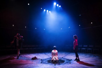  Zeppo (Guy Rhys) paces around an heptagonal shaped stage, which slopes slightly into a drain in the centre where a young girl is sat in a white dress wearing a full head prosthetic C'thulu mask. A large array of lights case moonlight from above. Ollie (Nadia Clifford) stands to the right of the image in a red jacket watching Zeppo pace.  