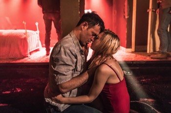  Eddie (Adam Rothenberg) and May (Lydia Wilson) both hold each other and press their heads together whilst kneeling on the floor. The motel room behind them glows red. 