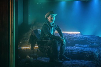  The Old Man (Joe McGann) sits in camping chair on a mound of mud. There are illuminated white fluorescent tubes laying in the mud. The Old Man is lit strongly in a teal colour with a strong blue backlight. 