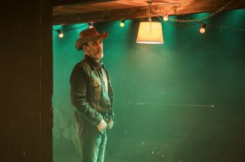  The Old Man (Joe McGann) stands inside the motel room. There's a green backlight shining into the room from the missing back wall of the motel room. A lit lampshade hangs just beside the Old Man's head. 