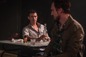  Eddie (Adam Rothenberg) sits at a table drinking alcohol with Martin (Luke Neal). 