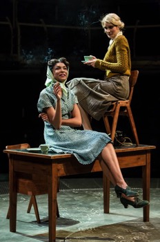  Helen Giniver (Kelly Hotten) sits on a chair on top of a desk, with Viv Pearce (Thalissa Teixeira) sits on the edge of the desk. Both are wearing 40s office clothing. They have cups of tea and are smoking. The lighting suggests they're outside on a bright but cold day. 