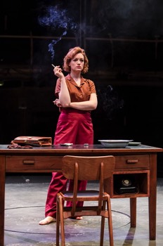  Julia Standing (Lucy Briggs-Owen) stands behind a writing desk, her arms folded smoking a cigarette. She wears red trousers and a dark tan blouse. 