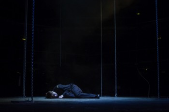 Kay Langrish (Jodie McNee) lays collapsed in the centre of the stage. There is a cold light from above creating a pool around her. She is surrounded by chains hanging from above. 