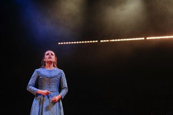  Zoe (Iola Evans) stands in her pale blue dress. She looks up worried. There is a long line of warm glowing lights behind her. 