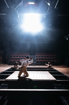  George (Ken Nwosu) kneels centre stage swinging a dagger through the air towards an imaginary character during a fight scene with his double-rolled character, M'Closky. He's on a small platform over a deep hole within the stage. Two women are screaming at the back of the stage stood within the pit and a bright blinding light flashes overhead. 