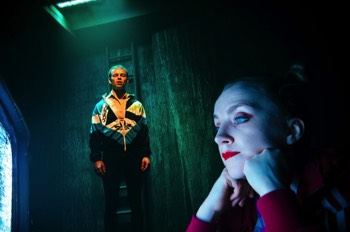 Runt (Evanna Lynch) lays on the floor, propping up her head on her hands, staring into a tv screen showing static, the glow on her face, with bright red lipstick. Behind her, Pig (Colin Campbell) stands looking at her. Pig wears a 90s Adidas zip up jacket over a yellow polo shirt. He's isolated, lit in green from above. There's a ladder on the wall just behind him up to the ceiling.
