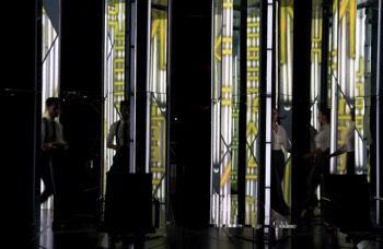 Seth (Tom Riley) and Jenny (Hayley Atwell) walking between mirrored toblerone shaped panels. Projection from behind shows white and yellow neon reminiscent of a Tokyo city billboard reflected between the panels.