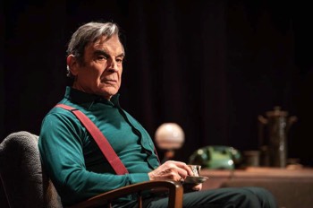  Harry (David Suchet) sits in profile in an armchair with a cup of tea. He wears a deep green long sleeved shirt with primary red braces. He looks into middle distance sligtly towards the camera. He looks unamused. 