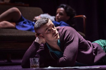  Bill (Russell Tovey) lays on the floor propped up on one arm. He's reading a magazine laid on the floor in front of him with a tumbler of whiskey. In the background, Stella (Hayley Squires) is laying on a sofa with a white cat on her chest. 