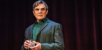  Harry (David Suchet) stands holding a empty whiskey glass. He is stood in front of purple curtains wearing a dark grey suit jacket over a green shirt. He looks displeased. 