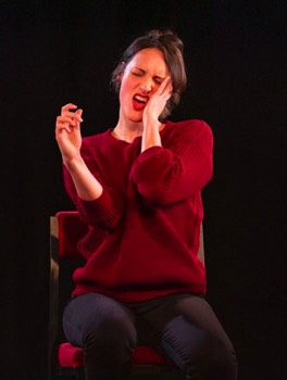 Phoebe Waller-Bridge as Fleabag. She is sat on a high stool. She pushes her bent elbows together miming taking a topless selfie. She is wearing a red knitted jumper and black tight jeans. Her left hand is on her face and attempting to pull a sexy face.