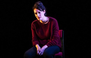 Phoebe Waller-Bridge as Fleabag. She is sat on a high stool. Her hands are in her lap and she's leaning forward looking unimpressed. The light across her looks like it's night time, but inside with warm lamps on, like in a bedroom.