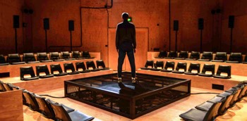  Ivan (Alex Austin) stands in the centre of the square stage. His back is to the camera and he is silhouetted in a cold white light. The square stage platform is made of black metal mesh with a corner towards the camera. The stage is surrounded on four sides by parallel seating banks constructed out of untreated wooden boards with black plastic seats bolted to it. Behind the each bank of seats are 3 speakers on poles. The walls are made of the same wood panelling. 