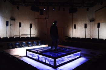  Ivan (Alex Austin) is stood in the centre of the square stage. The black mesh stage platform is underlit brightly with cold white light. Coiled cables are visible on the floor under the mesh. A bright white light is glowing out from behind the tall speakers behind the audience seating banks, so the wood panels of the walls are lit brightly, but Ivan in the centre is completely in the dark. 