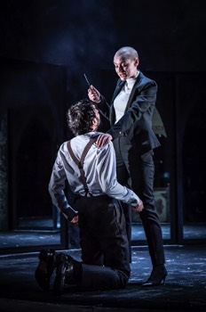  Ratcliffe (Heledd Gwynn) stands over Clarence (Tom Kanji) who kneels in front of her. She holds a knife in her right hand with her left holding Catesby's shoulder. 