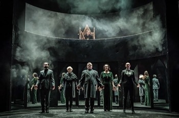  The full company stand spread apart across the stage for Richard III's (Tom Mothersdale) coronation. He is visible on a throne on a balcony above at the back of the stage. Behind the company on the stage floor are the arched mirror panels which give the effect of a larger crowd. The light on them all is green-tinged, like an overcast day. Stage smoke mist rises at the sides and catches the light. 