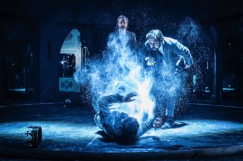  Richard III (Tom Mothersdale) lays on the floor shielding himself from the ghosts of Clarence (Tom Kanji) and Henry (John Sackville) who stand over him. Richard is a tight white pool of light. Clarence blows white dust over Richard creating a large plume over him which Richard tries to waft away. Henry looks on directly behind. 
