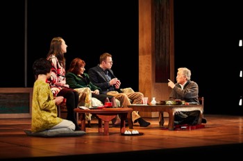 Missing People Production Image. The full company sit at a Japanese low dining table eating snacks. The room is abstract, with a warm wood floor and some wooden columns suggesting pillars. 