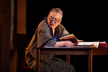 Missing People Production Image. Masaru Nakamura (Yutaka Oda) sits at his desk reading, looking at someone to his right who is out of shot. The room is dark, but warm.