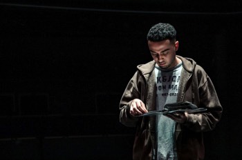 Louis (Ali Barouti) goes through his CD folder. He's in a spot light, looking like he's outside on a cold day. He's wearing an open zip-up brown hoodie with a grey t-shirt underneath. The rest of the space is black.