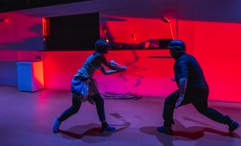 A Fight Against Production Image. The Girl (Pía Laborde-Noguez) swings her handbag at the Bouncer (Joseph Balderama) who is wearing a stab vest and military helmet. The back wall of the white wash set is lit in bright red light from inside the off-stage nightclub.