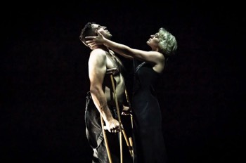 Julie (Heledd Gwynn) holds John's head (Tim Pritchett) with her arms outstretched. John is stood on wooden crutches. He is topless with braces hanging to the sides of his brown corded period trousers. Julie is wearing a black night gown. They are lit tightly in a white spotlight, the rest of the space is completely dark.