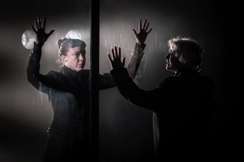Julie (Heledd Gwynn) reaches towards a glass wall with Christine (Catrin Aaron) behind, her arms up and hands pressed against the glass. There are lights shining straight outward behind Christine. Julie is silhouetted, and the rest of the space is completely dark.