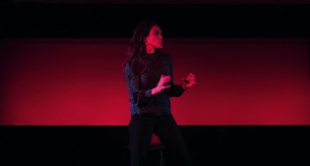She (Emily Bruni) is standing, her arms up defensively. She looks angry, turned slightly to her left. There is a letter box shaped screen behind her lit entirely in red from below. She is lit in red from in front.