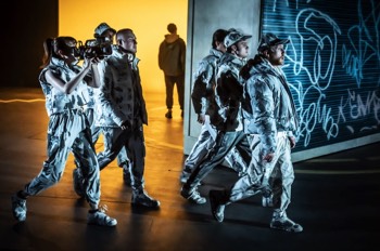 The ensemble dressed in white and grey camouflage jackets walk together past a graffiti covered roller shutter.  Someone stands in the background silhouetted against an orange lit cyclorama.