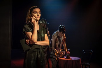 Mel (Isobel Fairchild) stands on the phone, dimly lit as a table of tapas is set up behind her.