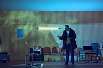 Jitney Production Image. Becker (Wil Johnson) stands centre stage, the wood panelled office is washed in a cold bluish moonlight. He's surrounded by wisps of smoke and holds out a dollar bill to nobody.