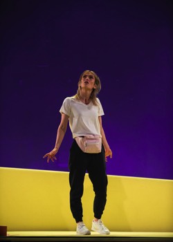 Purple Snowflakes and Titty Wanks Production Image. Saorise (Sarah Hanley) stood in the middle of the yellow carpeted stage. Purple lights shine up from behind the set.