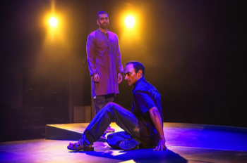 Zafar (Esh Alladi) sits on the floor with his hands behind him. Bilal (Waleed Akhtar) stands in the background wearing a light red kurta. Two warm lights shine behind Bilal towards Zafar, who is bathed in deep blue light.