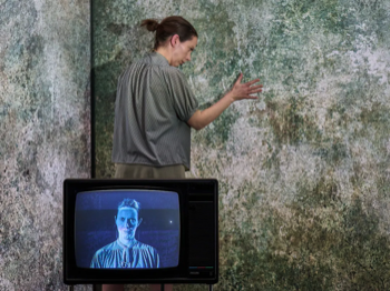 The woman (Kate O'Flynn) stood against a mould covered wall reaching her hand out to touch it. An old television set is in the foreground with an image of the woman on the screen.