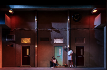 Wide shot of the stage, it's night time with large sodium street lamps attached to the walls of the concrete tower block. Moonlight skims across the building front whilst Jamie (Rilwan Abiola Owokoniran) and Ste (Raphael Akuwudike) hang out outside Jamie's flat.
