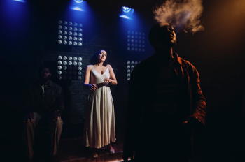Christina (Aimee Powell) in a white silk dress with Moses (Gamba Cole) smoking in shadows in the foreground.