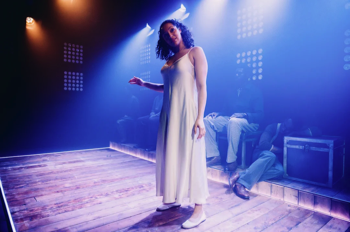 Christina (Aimee Powell) in a white silk dress placing her hand on the head of an imaginary child, strongly backlit.