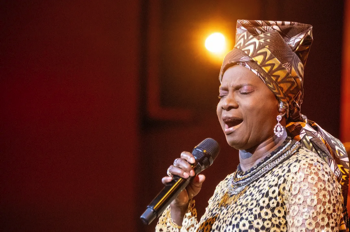 Angélique Kidjo singing wearing a gold head dress and gold patterned dress