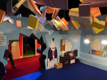 The 100 Story Hotel Reception with walls painted like the sky and books floating across the ceiling. Clive the polar bear concierge is stood beside a large hole in the wall.